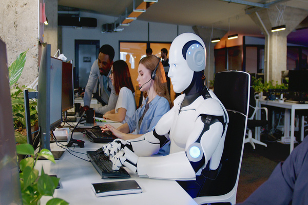 A robot sat at a desk with two women and a man working at a computer so signify machine learning and AI within IT support