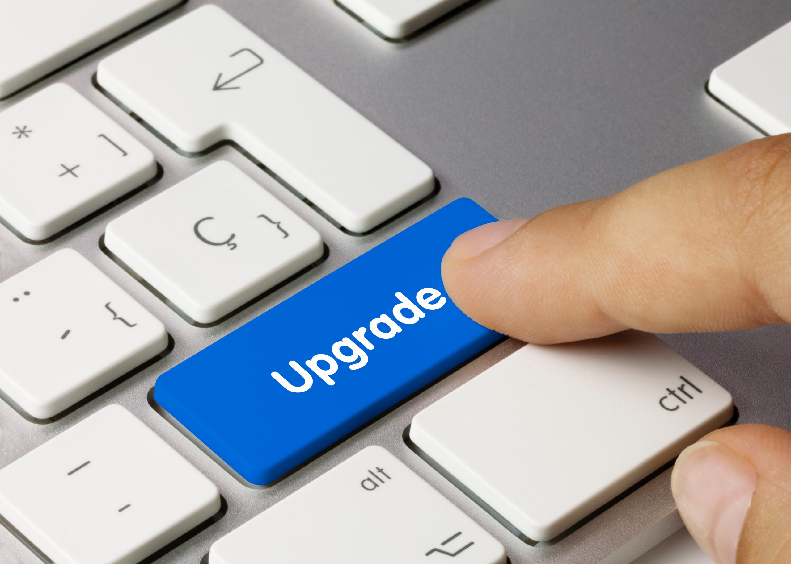 Grey computer keyboard with the shift key changed to be blue, with the word "upgrade" written on it. A finger is pointed over the button as if to press it.