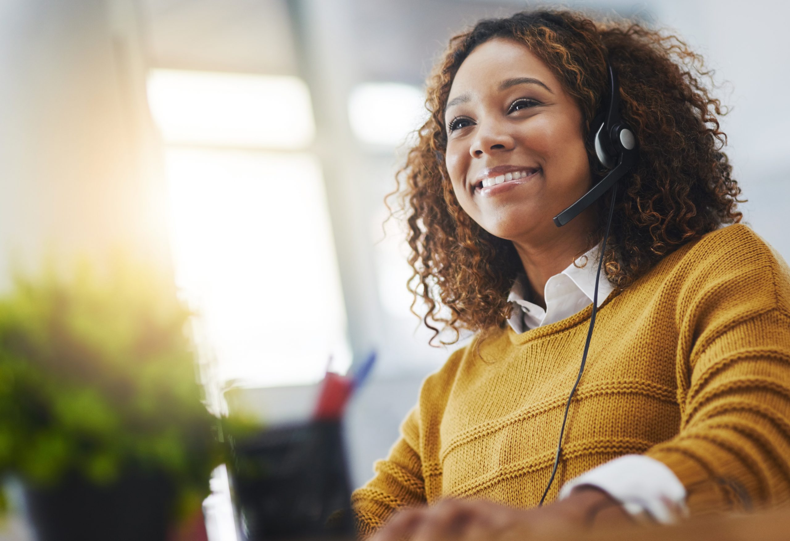 Young Black woman wearing a yellow jumper working in a call centre talking on a VoIP headset in front of a laptop
