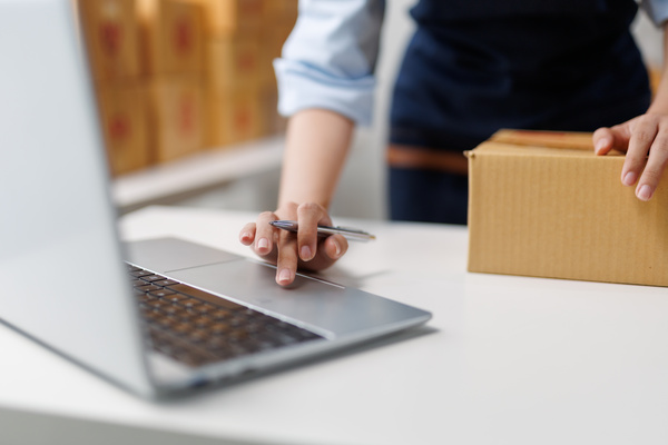 Person using a laptop while holding a cardboard box, likely preparing an online order in a workspace.