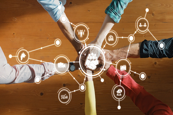 Top‑down view of several hands joined in the centre with digital network icons overlaid, representing teamwork and interconnected systems