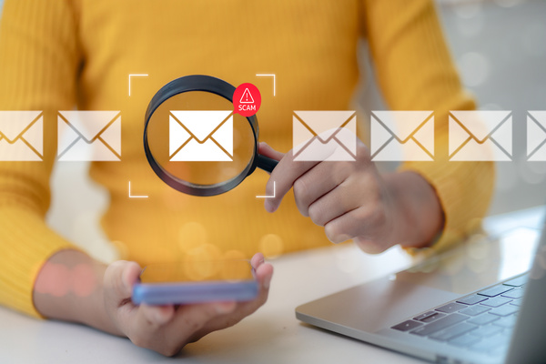 Person holding a smartphone next to a laptop with a magnifying glass highlighting an email icon marked as a scam.